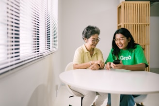 Two women are seated at a white table in a bright room. They appear engaged in a friendly interaction, with one woman showing something on her phone to the other. The room has white walls with a window covered by blinds, allowing natural light to illuminate the scene.