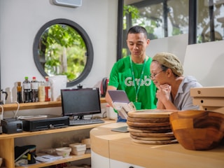 In a well-lit room with a circular window, two people focus on a screen. One person is wearing a green shirt and assisting the other, who is sitting at a counter with a thoughtful expression. Various bottles, a computer monitor, and wooden plates are on the counter, adding to the room's modern and organized ambiance.