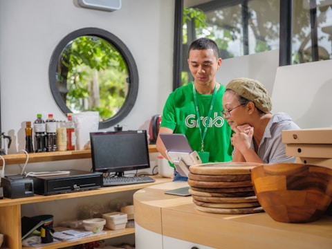 In a well-lit room with a circular window, two people focus on a screen. One person is wearing a green shirt and assisting the other, who is sitting at a counter with a thoughtful expression. Various bottles, a computer monitor, and wooden plates are on the counter, adding to the room's modern and organized ambiance.