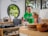 In a well-lit room with a circular window, two people focus on a screen. One person is wearing a green shirt and assisting the other, who is sitting at a counter with a thoughtful expression. Various bottles, a computer monitor, and wooden plates are on the counter, adding to the room's modern and organized ambiance.
