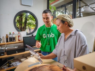 Two people are standing indoors. One person, wearing a green shirt with a logo, is smiling and assisting another person who is looking at a tablet device. The setting is a room with shelves in the background holding various bottles and a computer monitor. A large window allows natural light to illuminate the space.