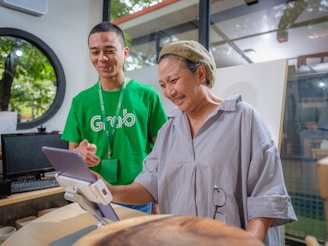 A man in a green shirt with a logo assists a woman in front of a tablet mounted on a stand. The setting appears to be indoors with large windows revealing trees outside. The man is smiling while the woman seems engaged and amused by what she is seeing on the tablet.