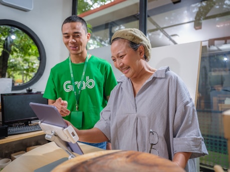 A man in a green shirt with a logo assists a woman in front of a tablet mounted on a stand. The setting appears to be indoors with large windows revealing trees outside. The man is smiling while the woman seems engaged and amused by what she is seeing on the tablet.