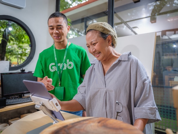 A man in a green shirt with a logo assists a woman in front of a tablet mounted on a stand. The setting appears to be indoors with large windows revealing trees outside. The man is smiling while the woman seems engaged and amused by what she is seeing on the tablet.