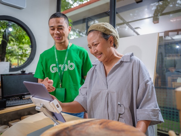 A man in a green shirt with a logo assists a woman in front of a tablet mounted on a stand. The setting appears to be indoors with large windows revealing trees outside. The man is smiling while the woman seems engaged and amused by what she is seeing on the tablet.