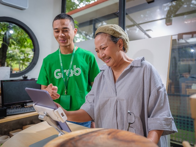 A man in a green shirt with a logo assists a woman in front of a tablet mounted on a stand. The setting appears to be indoors with large windows revealing trees outside. The man is smiling while the woman seems engaged and amused by what she is seeing on the tablet.