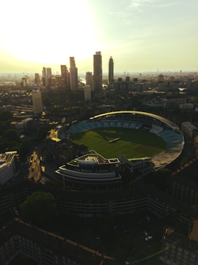 Aerial view of a cricket stadium surrounded by urban infrastructure, featuring a modern skyline with high-rise buildings in the background. The sun is setting or rising, casting a warm glow over the scene.
