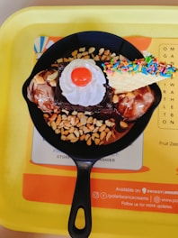 A dessert presented in a black skillet, including a piece of chocolate brownie, adorned with whipped cream, a cherry on top, two scoops of ice cream, crushed peanuts, and a wafer cone with colorful sprinkles, resting on a yellow tray.