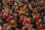 A large group of children of various ages are gathered closely together, with many looking directly at the camera. Some children are smiling, others are making playful hand gestures, and they are dressed in colorful clothing.