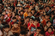 A large group of children of various ages are gathered closely together, with many looking directly at the camera. Some children are smiling, others are making playful hand gestures, and they are dressed in colorful clothing.