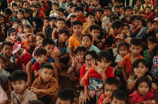 A large group of children of various ages are gathered closely together, with many looking directly at the camera. Some children are smiling, others are making playful hand gestures, and they are dressed in colorful clothing.