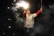 A person is joyfully holding sparklers against a dark background. Wisps of smoke and bright sparks surround the figure, who is wearing a shiny, reflective jacket and appears to be celebrating with a broad smile.