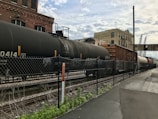 A train with several freight cars is situated on the tracks adjacent to an industrial building, with a chain-link fence in the foreground. The freight cars include tankers and boxcars, and the setting features overcast skies and a factory-like backdrop. The ground alongside the tracks consists of asphalt and patches of grass.