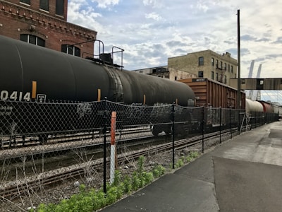 A train with several freight cars is situated on the tracks adjacent to an industrial building, with a chain-link fence in the foreground. The freight cars include tankers and boxcars, and the setting features overcast skies and a factory-like backdrop. The ground alongside the tracks consists of asphalt and patches of grass.
