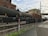 A train with several freight cars is situated on the tracks adjacent to an industrial building, with a chain-link fence in the foreground. The freight cars include tankers and boxcars, and the setting features overcast skies and a factory-like backdrop. The ground alongside the tracks consists of asphalt and patches of grass.
