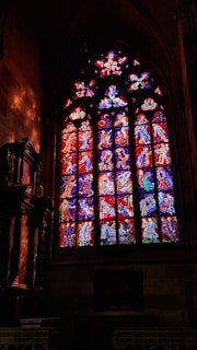 A large stained glass window depicting various biblical scenes with intricate details and vibrant colors. The craftsmanship highlights figures and narratives from religious texts, illuminated by ambient light.