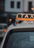 A close-up of the roof of a taxi with an illuminated sign displaying the word 'TAXI.' The background shows blurred outlines of buildings and lights.