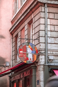 A corner building with a vintage sign of a bakery depicting a baker holding a paddle with a loaf of bread. The building's exterior features stone bricks and muted pink and brown tones. The bakery's name, 'Boulangerie du Palais', is visible above the windows.