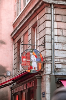 A corner building with a vintage sign of a bakery depicting a baker holding a paddle with a loaf of bread. The building's exterior features stone bricks and muted pink and brown tones. The bakery's name, 'Boulangerie du Palais', is visible above the windows.