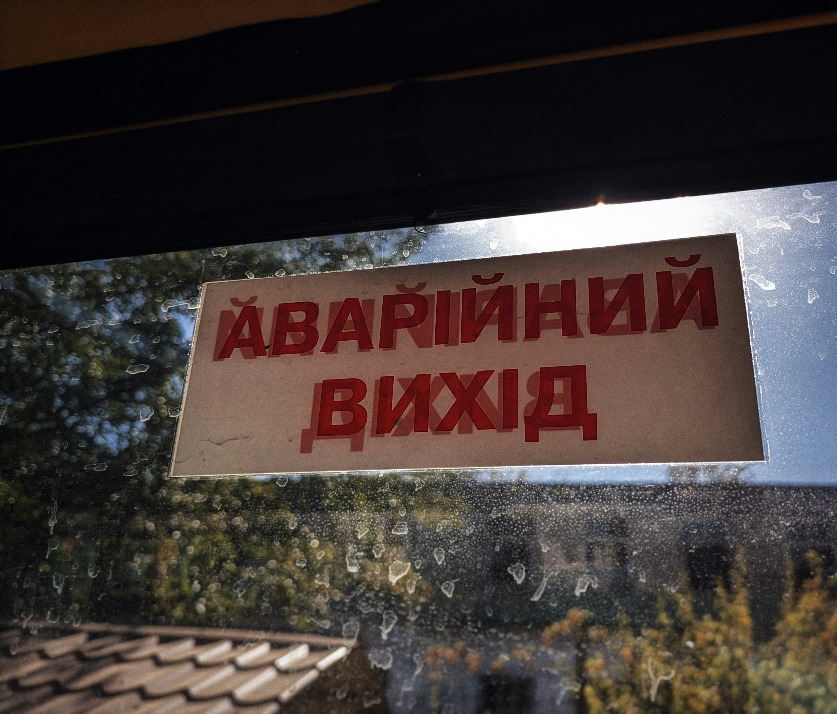 A sign with red text in a non-Latin script is displayed on a window with a view of trees and a building. The windowpane is speckled with dirt, and sunlight is visible.