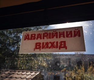 A sign with red text in a non-Latin script is displayed on a window with a view of trees and a building. The windowpane is speckled with dirt, and sunlight is visible.