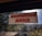 A sign with red text in a non-Latin script is displayed on a window with a view of trees and a building. The windowpane is speckled with dirt, and sunlight is visible.