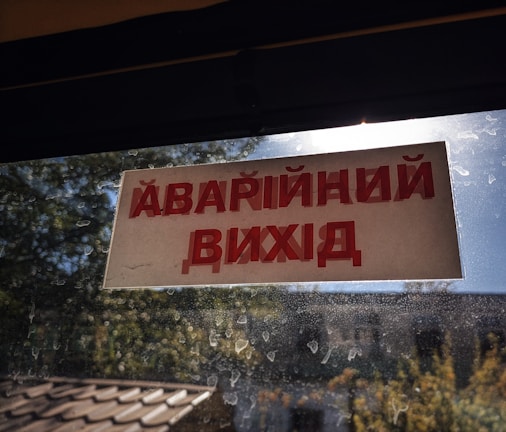 A sign with red text in a non-Latin script is displayed on a window with a view of trees and a building. The windowpane is speckled with dirt, and sunlight is visible.
