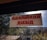 A sign with red text in a non-Latin script is displayed on a window with a view of trees and a building. The windowpane is speckled with dirt, and sunlight is visible.