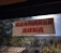 A sign with red text in a non-Latin script is displayed on a window with a view of trees and a building. The windowpane is speckled with dirt, and sunlight is visible.