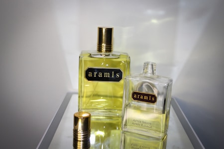 Two bottles of Aramis fragrance are displayed on a reflective surface. The larger bottle on the left contains a yellow liquid and has a golden cap, while the smaller bottle on the right is partially open. Both bottles feature the Aramis label prominently.