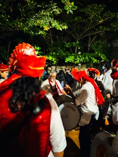A group of people are gathered outdoors at night, with several individuals wearing bright red traditional headgear and playing large drums. The surrounding trees are lush and vibrant, partially illuminated by artificial lighting. The scene is lively, suggesting a cultural or festive celebration.