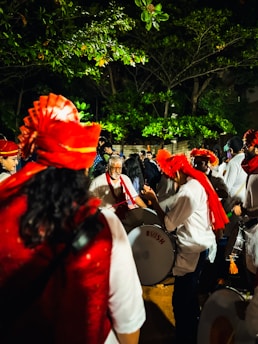 A group of people are gathered outdoors at night, with several individuals wearing bright red traditional headgear and playing large drums. The surrounding trees are lush and vibrant, partially illuminated by artificial lighting. The scene is lively, suggesting a cultural or festive celebration.