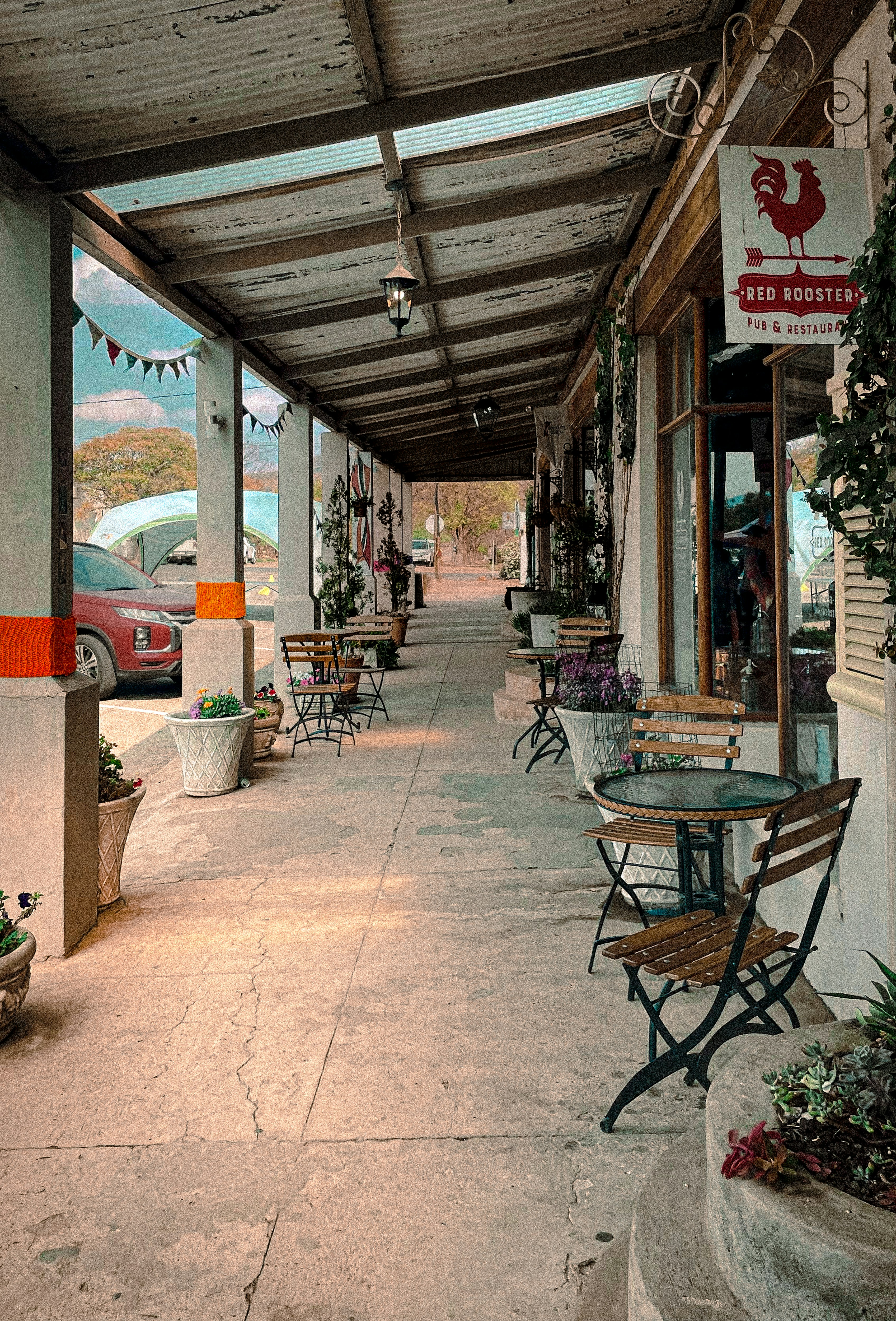 A cozy outdoor café setting features several wooden tables with chairs under a sheltered walkway. Potted plants line the edges, adding greenery to the concrete paths. A sign for 'Red Rooster Pub & Restaurant' hangs above one of the doors. A car is visible parked on the side, while a string of colorful flags is hung between the columns.