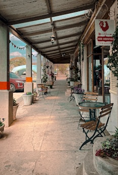 A cozy outdoor café setting features several wooden tables with chairs under a sheltered walkway. Potted plants line the edges, adding greenery to the concrete paths. A sign for 'Red Rooster Pub & Restaurant' hangs above one of the doors. A car is visible parked on the side, while a string of colorful flags is hung between the columns.