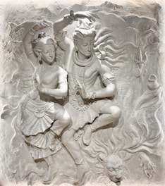 A detailed relief sculpture depicting two figures in a dynamic dance pose, adorned with elaborate jewelry and traditional attire. The background features swirling motifs and a lion's head figure, adding drama and movement to the scene.