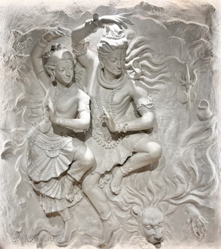 A detailed relief sculpture depicting two figures in a dynamic dance pose, adorned with elaborate jewelry and traditional attire. The background features swirling motifs and a lion's head figure, adding drama and movement to the scene.