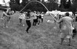 A group of people is outdoors on a grassy field, participating in a jump rope activity. Several adults are turning a long rope while children take turns jumping. Tents and trees are visible in the background, suggesting a festival or community event.