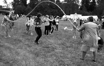 A group of people is outdoors on a grassy field, participating in a jump rope activity. Several adults are turning a long rope while children take turns jumping. Tents and trees are visible in the background, suggesting a festival or community event.