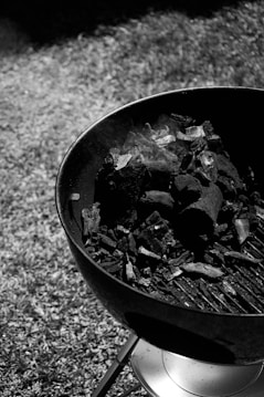 A round charcoal grill with dark coals and some smoke rising from it, sitting on a metal stand on a grassy surface. The grill is open and appears to be ready for use.