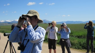 Several people are birdwatching in an open grassy area with mountains and a clear blue sky in the background. They are using binoculars and cameras to observe their surroundings. One person is wearing a wide-brimmed hat and another has a camera with a large lens mounted on a tripod.