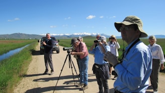 A group of people are standing on a dirt road in a rural area with a scenic background. They are engaged in bird watching or nature observation; several are using binoculars and one person has a camera mounted on a tripod. The scene features a clear blue sky, distant snow-capped mountains, and a vehicle parked alongside the road. There is green grass beside the road and a small water body.