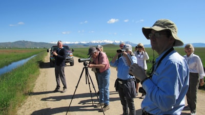 A group of people are standing on a dirt road in a rural area with a scenic background. They are engaged in bird watching or nature observation; several are using binoculars and one person has a camera mounted on a tripod. The scene features a clear blue sky, distant snow-capped mountains, and a vehicle parked alongside the road. There is green grass beside the road and a small water body.