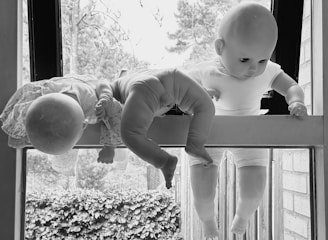 Three baby dolls are arranged with their heads and limbs hanging down a window sill, overlooking an outdoor scene with foliage in the background. The image is in black and white, giving it a dramatic and eerie feel. The dolls appear to be in various positions, some wearing clothing.