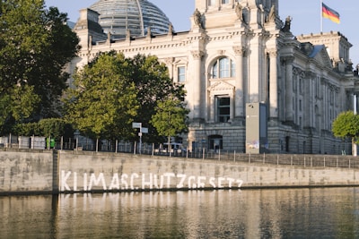 A historic and grand building with ornate architectural details stands prominently by a calm river. The structure features large windows and a dome, indicating a significant or governmental facility. A German flag is visible on top of the building. In front of the building, along the riverbank, the word 'Klimaschutzgesetz' is painted in white on the wall above the water. Lush green trees partially shade the area, and the weather appears clear and sunny.
