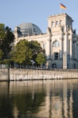 A large historic building with a prominent glass dome is seen in the background, adorned with an ornamental facade. The structure is flanked by lush green trees and a waterway in the foreground. A German flag flies atop the building. On the retaining wall along the waterway, the word 'KLIMASCHUTZGESETZ' is spray-painted in white.