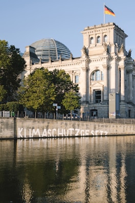 A large historic building with a prominent glass dome is seen in the background, adorned with an ornamental facade. The structure is flanked by lush green trees and a waterway in the foreground. A German flag flies atop the building. On the retaining wall along the waterway, the word 'KLIMASCHUTZGESETZ' is spray-painted in white.