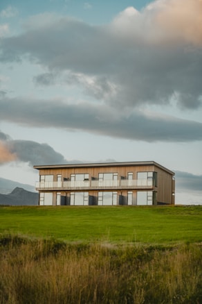 A modern two-story building with a sleek and minimalist design sits amidst a vast green field. Large windows line the front of the structure, offering expansive views of the surrounding landscape. The sky above is partly cloudy, with fluffy clouds creating a dramatic backdrop.