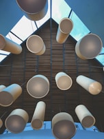 A structured architectural ceiling features large cylindrical light fixtures hanging in a symmetrical pattern. The ceiling has a geometric design with wooden panels and glass sections, through which a clear blue sky is visible.