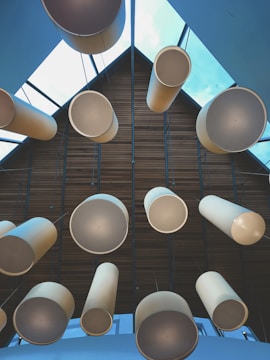 A structured architectural ceiling features large cylindrical light fixtures hanging in a symmetrical pattern. The ceiling has a geometric design with wooden panels and glass sections, through which a clear blue sky is visible.