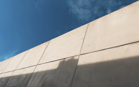 A large concrete wall is visible under a clear blue sky. The wall is segmented into panels and casts a shadow on its surface.